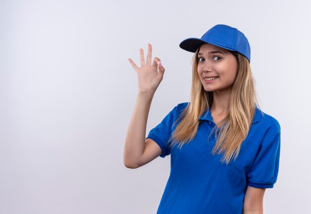 Ragazza di consegna giovane interessata che indossa l'uniforme blu e cappuccio che mostra il gesto okey isolato su fondo bianco con lo spazio della copia