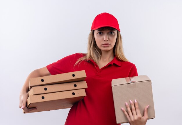 Ragazza di consegna giovane interessata che porta l'uniforme rossa e la scatola della tenuta del cappuccio e le scatole per pizza isolate sulla parete bianca