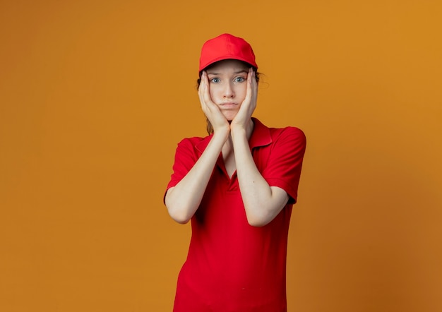 Ragazza di consegna graziosa giovane confusa in uniforme rossa e cappuccio che mette le mani sul viso che guarda l'obbiettivo isolato su priorità bassa arancione con lo spazio della copia