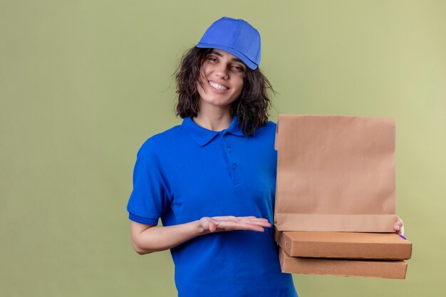 Ragazza di consegna in uniforme blu che tiene scatole per pizza e pacchetto di carta che presenta con il braccio della mano che sorride allegramente sorridente sopra lo spazio verde isolato