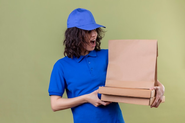 Ragazza di consegna in uniforme blu che tiene scatole per pizza e pacchetto di carta che sembra sorpreso e felice in piedi sopra lo spazio verde