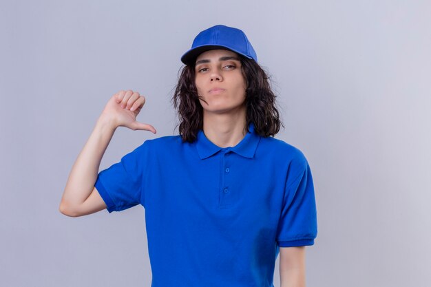 Ragazza di consegna in uniforme blu e cappuccio che indica il pollice su se stessa con espressione seria e sicura in piedi sul bianco isolato