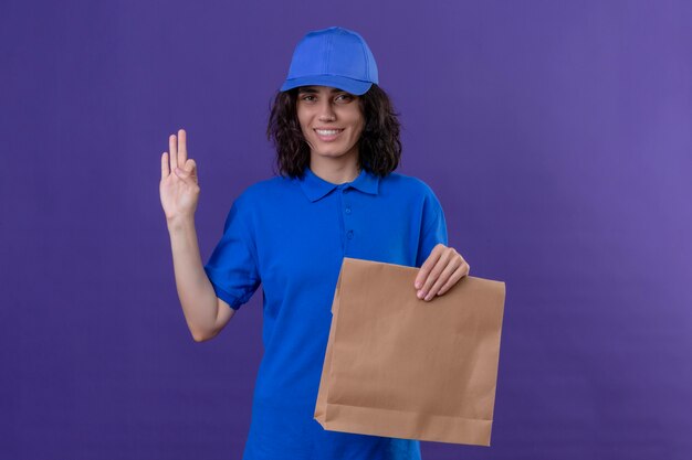 Ragazza di consegna in uniforme blu e cappuccio che tiene il pacchetto di carta che sembra sicuro e felice facendo segno giusto che sta sulla porpora isolata