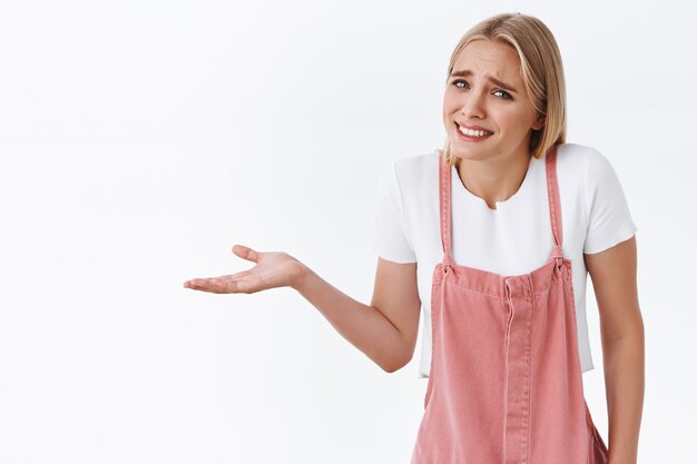 Ragazza di fronte a una situazione scomoda, alzando le spalle e allargando una mano lateralmente, sorridendo scioccamente e le sopracciglia solcate sembrano deluse e imbarazzate, in piedi sfondo bianco