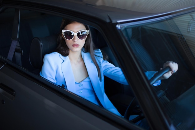 Ragazza di moda che guida un'auto in un vestito blu. Donna alla moda che si siede in macchina e che tiene il volante. Sparato attraverso il finestrino laterale.