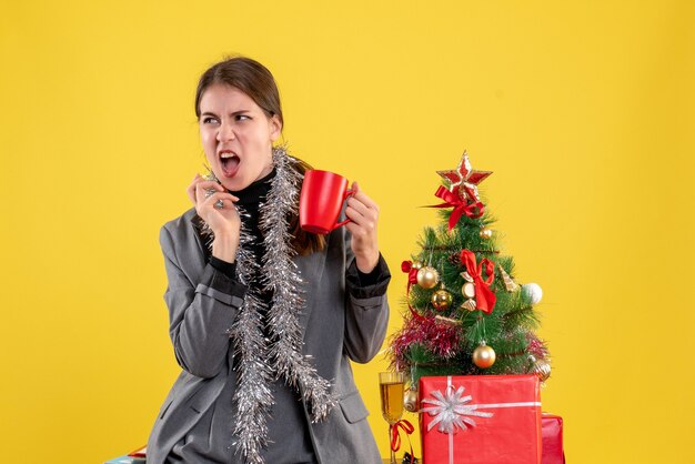 Ragazza di vista frontale con la bocca aperta che tiene tazza rossa vicino all'albero di Natale e cocktail dei regali