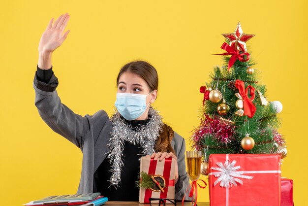 Ragazza di vista frontale con mascherina medica che si siede al tavolo salutando qualcuno albero di Natale e regali cocktail