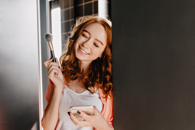 Ragazza di zenzero di risata che fa trucco. Ritratto dell'interno della giovane donna accattivante che tiene il pennello cosmetico.