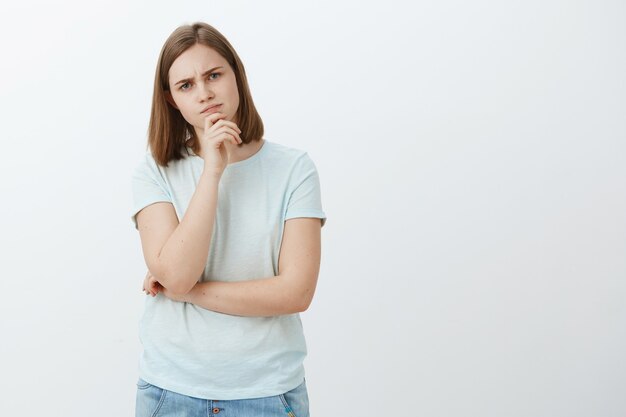 Ragazza difficile da impressionare. Studente femminile caucasico attraente premuroso intelligente e curioso che tiene la mano sul mento pensando accigliato mentre fa impressione in piedi e prende una decisione sul muro grigio