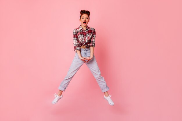 Ragazza divertente del pinup in blue jeans che salta e che guarda l'obbiettivo. Vista integrale della giovane donna in camicia a scacchi che balla sullo spazio rosa.