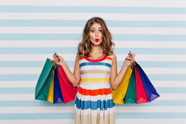 Ragazza divertente in vestito a strisce in posa con l'espressione del viso baciante dopo lo shopping. Affascinante giovane donna con i capelli ricci che tengono i sacchetti dalla boutique.