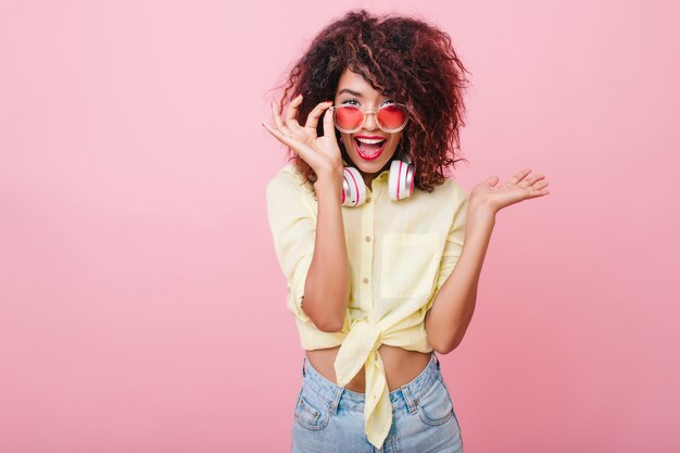 Ragazza eccitata con cuffie colorate in posa con piacere ed esprimere veramente emozioni. Signora romantica con un sorriso sorpreso che tiene i suoi occhiali da sole rosa.