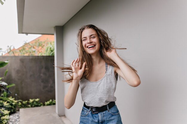 Ragazza eccitata in abiti alla moda ballando davanti al muro bianco. Colpo all'aperto di signora castana emotiva in jeans godendo.