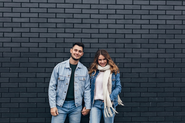 Ragazza eccitata in abito di jeans alla moda che tengono le mani con il ragazzo. Coppie amorose sorridenti che stanno insieme sul muro di mattoni.