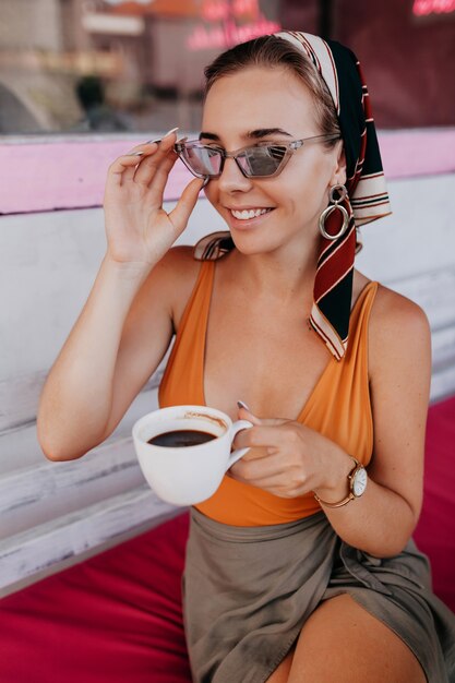 Ragazza emozionante con accessori alla moda che ride e che tiene gli occhiali da sole alla moda mentre beve il caffè davanti al ristorante