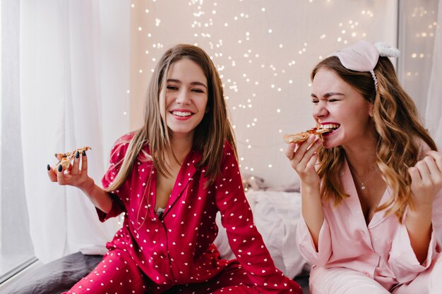 Ragazza entusiasta con l'acconciatura riccia che mangia pizza. Foto dell'interno della giovane donna caucasica felice in pigiama rosso che posa nella camera da letto con gli alimenti a rapida preparazione.
