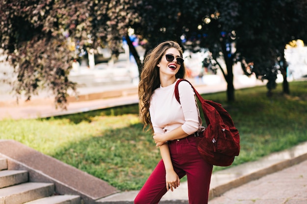 Ragazza espressiva con lunghi capelli ricci è in posa con la borsa vinosa nel parco della città. Indossa color marsala, occhiali da sole e buon umore.