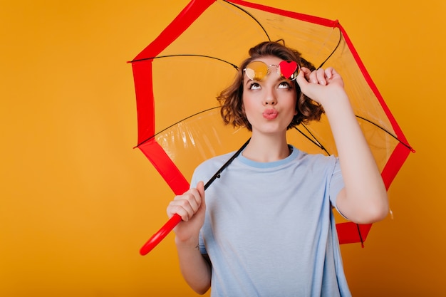 Ragazza estatica con l'espressione del viso divertente che tocca i suoi occhiali da sole e guardando il piccolo cuore. Foto dell'interno della giovane donna castana ispirata che posa con l'ombrello.