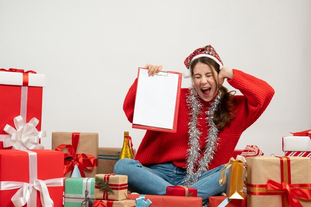 Ragazza euforica di vista frontale con i file della tenuta del cappello della Santa che si siedono intorno ai regali