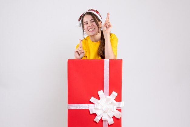 Ragazza euforica di vista frontale con il cappello della Santa che fa il segno di buona fortuna che sta dietro il grande regalo di natale