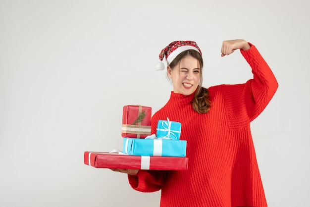 Ragazza euforica di vista frontale con il cappello della Santa che tiene i suoi regali di natale che mostrano il muscolo