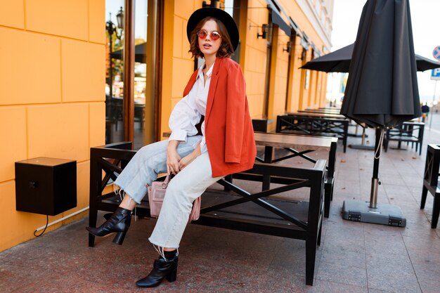 Ragazza europea sorridente in occhiali da sole arancioni carini, giacca e seduta black hat all'aperto. . Moda autunno