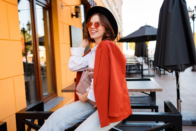 Ragazza europea sorridente in occhiali da sole arancioni carini, giacca e seduta black hat all'aperto.