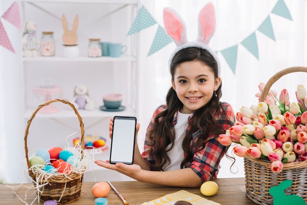 Ragazza felice che mostra telefono cellulare con cesto di uova di Pasqua e tulipani sul tavolo