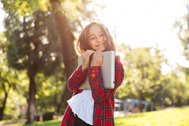 Ragazza felice che sta nel parco mentre abbracciando il suo computer portatile