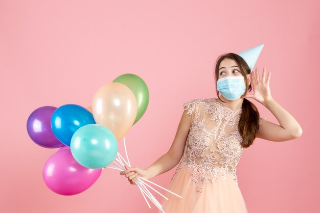 ragazza felice con cappello da festa e maschera medica ascoltando qualcosa mentre si tengono palloncini colorati sul rosa