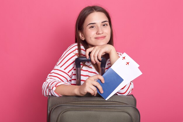 Ragazza felice con la valigia e il passaporto isolato sopra il muro rosa, ragazza dai capelli scuri in camicia a righe, indossa la maglietta casual a righe, essendo pronta a viaggiare.