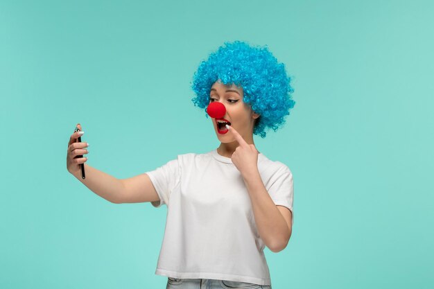 Ragazza felice del giorno dello sciocco di aprile di vista frontale che prende selfie che indica la bocca con i capelli blu del costume del pagliaccio del naso rosso