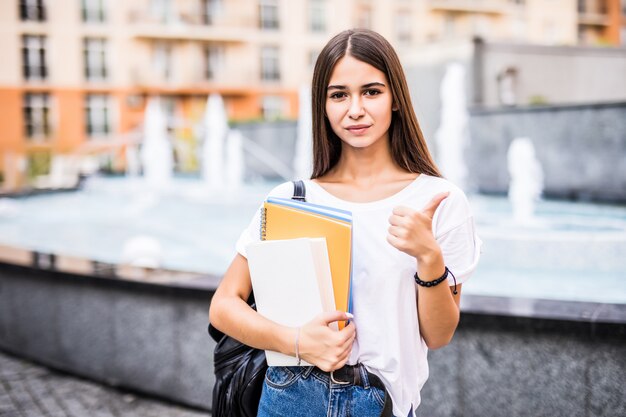 Ragazza felice dello studente che posa con i pollici su che ti esaminano nella via