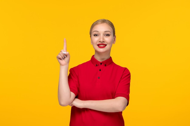 Ragazza felice di giorno della camicia rossa che sorride in camicia rossa e rossetto su fondo giallo che punta il dito verso l'alto