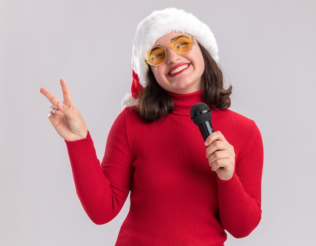 Ragazza felice e allegra in maglione rosso e cappello della santa con gli occhiali che tengono il microfono che sorride allegramente mostrando il segno di v in piedi sopra il muro bianco