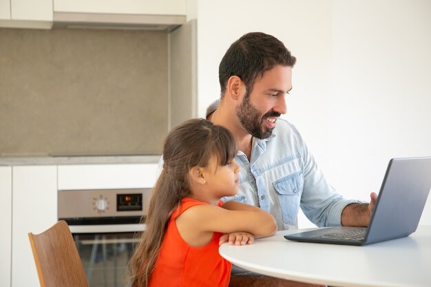 Ragazza felice e suo padre che utilizzano laptop per videochiamata, seduti a tavola, guardando il display e sorridendo.