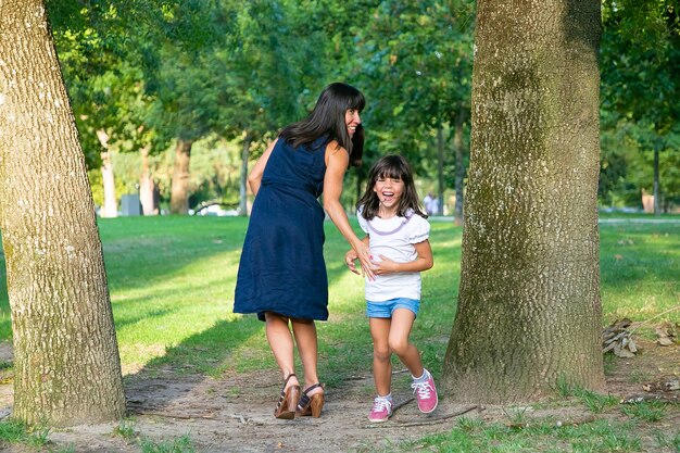 Ragazza felice eccitata che gioca giochi attivi con sua madre all'aperto, in piedi sugli alberi nel parco e ridendo. Lunghezza intera. Famiglia attività all'aperto e concetto di svago