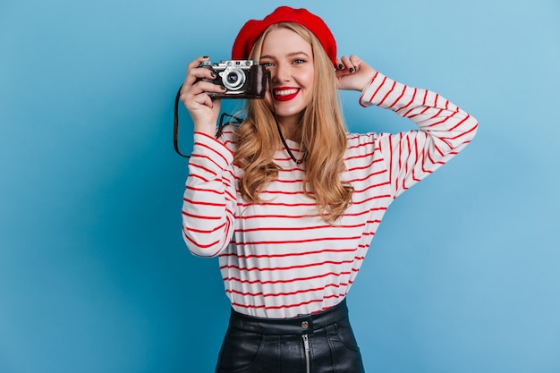 ragazza felice in camicia a righe tenendo la fotocamera. Modello femminile francese che cattura le foto sulla parete blu.