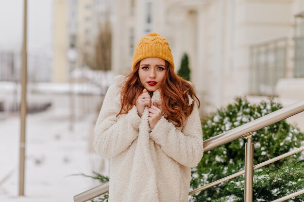 Ragazza felice in cappello giallo sveglio che gode dell'inverno. Signora interessata che trascorre la giornata di dicembre all'aperto.