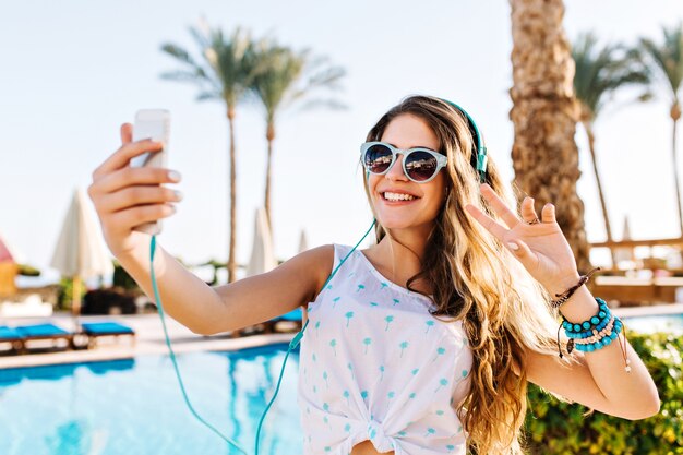 Ragazza felice in occhiali da sole con pelle abbronzata che fa selfie con segno di pace su sfondo di palme