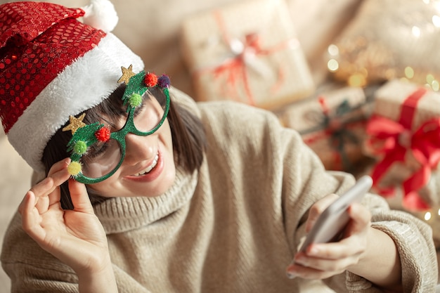 Ragazza felice in occhiali di Natale divertenti e cappello di Babbo Natale con un telefono in mano.