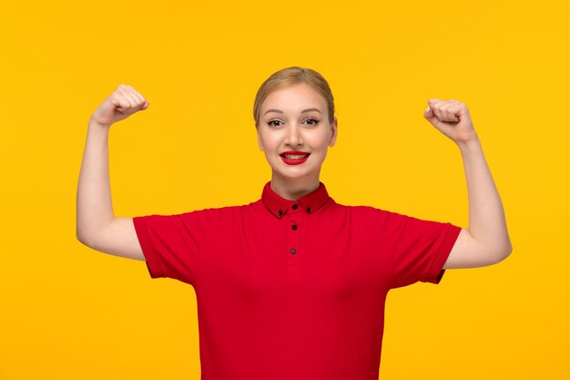 Ragazza felice sveglia di giorno della camicia rossa che mostra i suoi bicipiti in una camicia rossa su una priorità bassa gialla