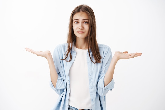 Ragazza femminile attraente ignara e all'oscuro con i capelli castani in camicetta blu sopra la maglietta bianca che scrolla le spalle con le mani alzate vicino alle spalle e sguardo dispiaciuto incerto