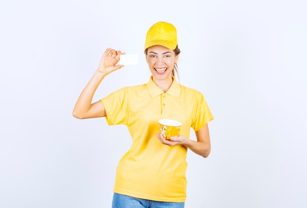 Ragazza femminile in uniforme gialla che consegna una tazza di noodle da asporto gialla e presenta il suo biglietto da visita al cliente.