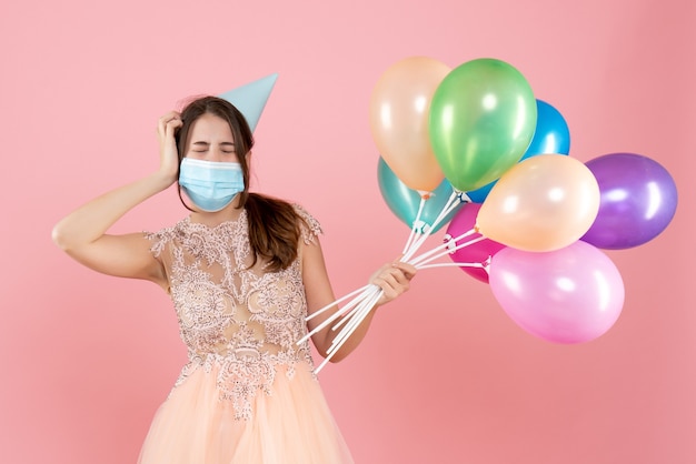 ragazza festaiola con cappello da festa e maschera medica chiudendo gli occhi tenendo palloncini colorati sul rosa