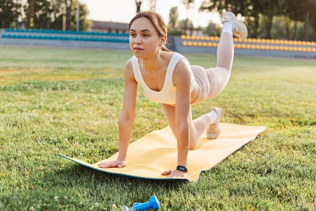 Ragazza fitness che fa allenamento per le gambe sul tappetino da yoga allo stadio all'aperto, donna in forma che indossa top bianco e leggins beige che si allenano da soli, assistenza sanitaria, stile di vita sano.