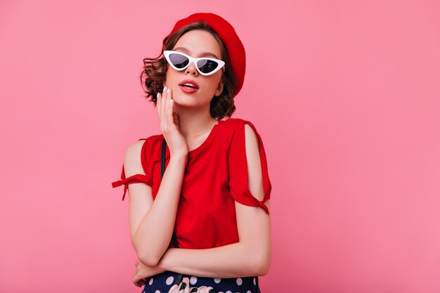 Ragazza francese ispirata con capelli ricci scuri in piedi. Meravigliosa donna caucasica in berretto rosso che guarda con interesse.