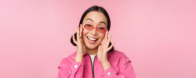 Ragazza giapponese moderna sveglia in occhiali da sole che sorride e che sembra felice in posa su sfondo rosa in abiti eleganti
