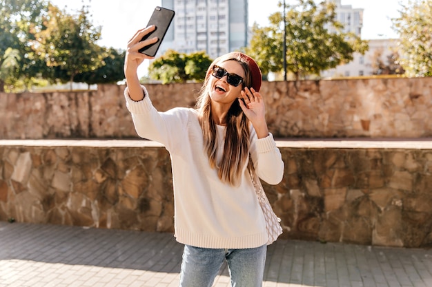 Ragazza gioconda in maglione bianco che fa selfie sulla strada. Donna ben vestita in berretto divertendosi in una buona giornata autunnale.