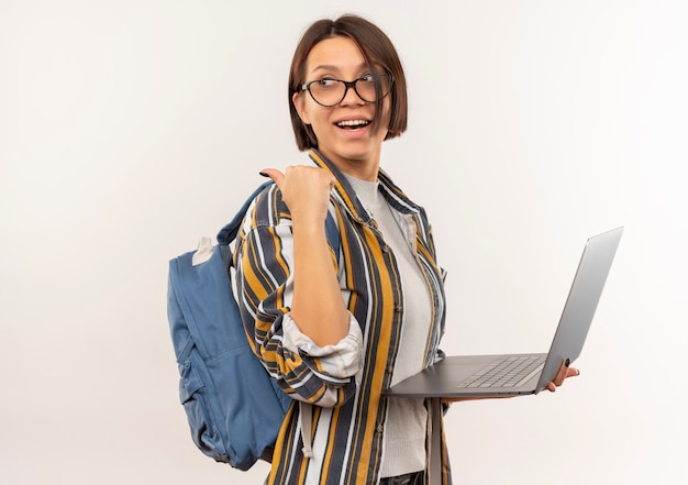 Ragazza gioiosa giovane studente con gli occhiali e borsa posteriore in piedi in vista di profilo tenendo il computer portatile che punta dietro isolato su bianco